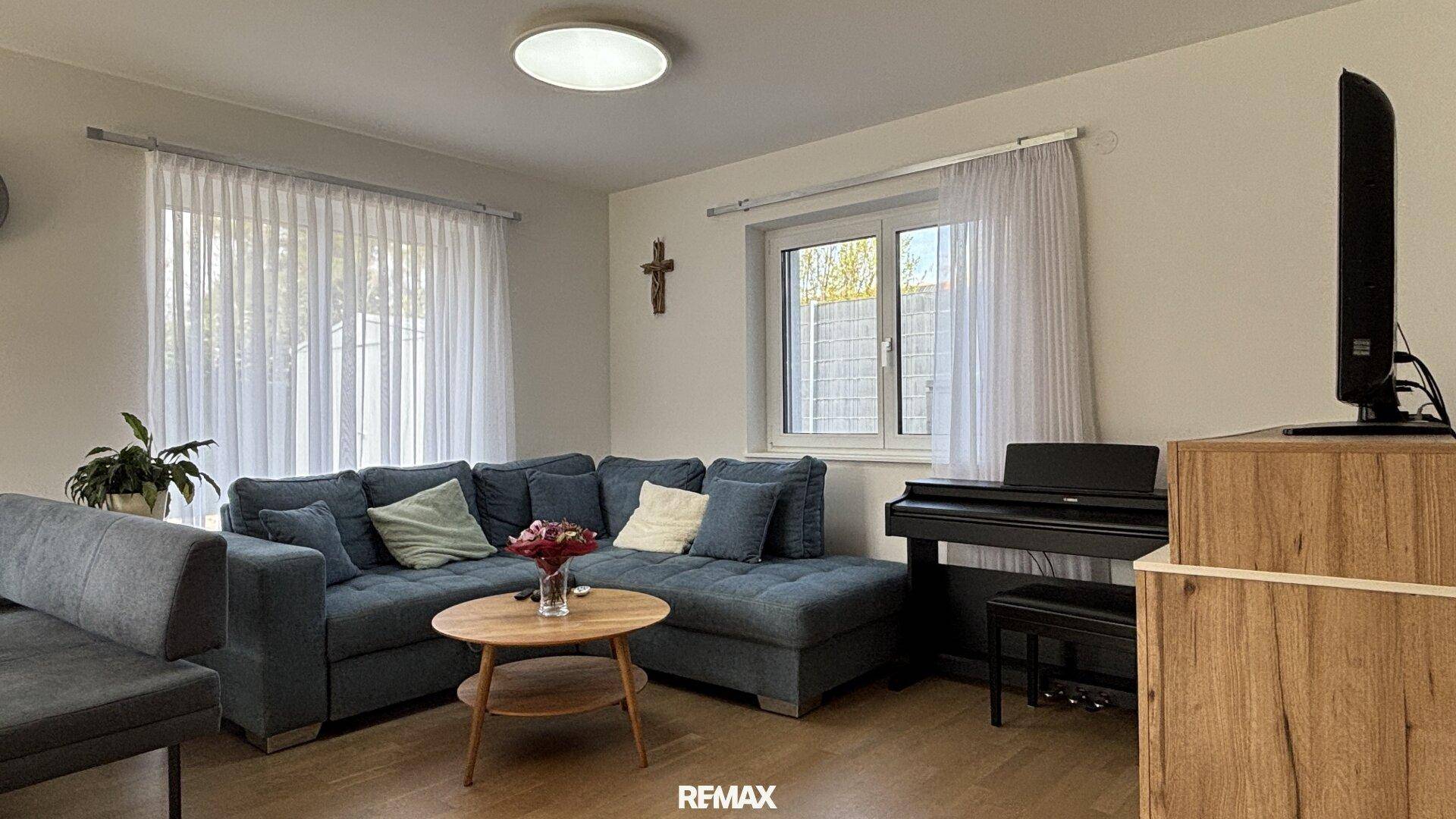 Gemütliches Wohnzimmer mit großem blauen Ecksofa, Holztisch und Klavier, hell durch Fenster.