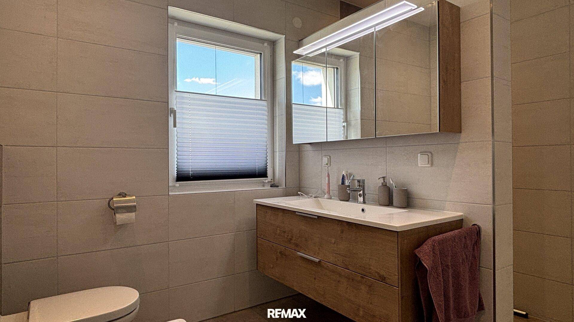 Modernes Badezimmer mit Waschtischunterschrank, Spiegelschrank und Fenster, in hellen Fliesen gehalten.