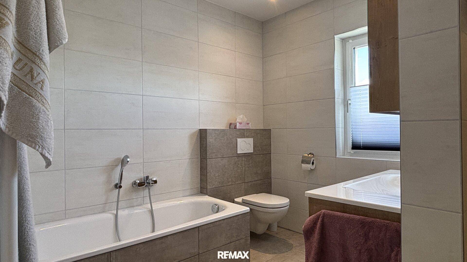 Helles Badezimmer mit hellgrauen Fliesen, Badewanne, wandhängendem WC und Fenster für Tageslicht.
