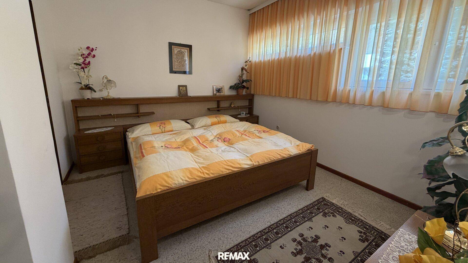 Schlafzimmer mit Doppelbett, Holzmöbeln und Teppichboden, hell durch großes Fenster.