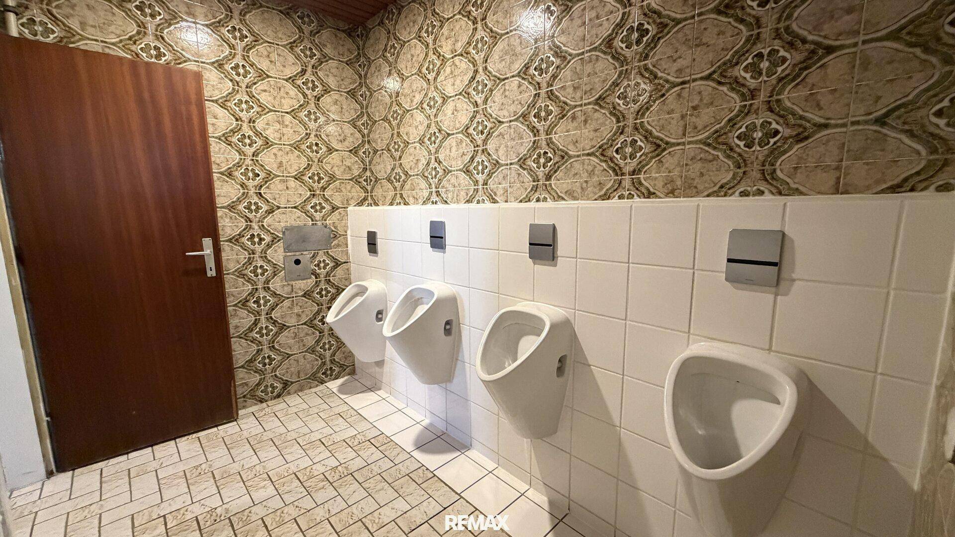 Herren-WC mit mehreren Urinalen, weißen Fliesen und gemusterten Wandfliesen.