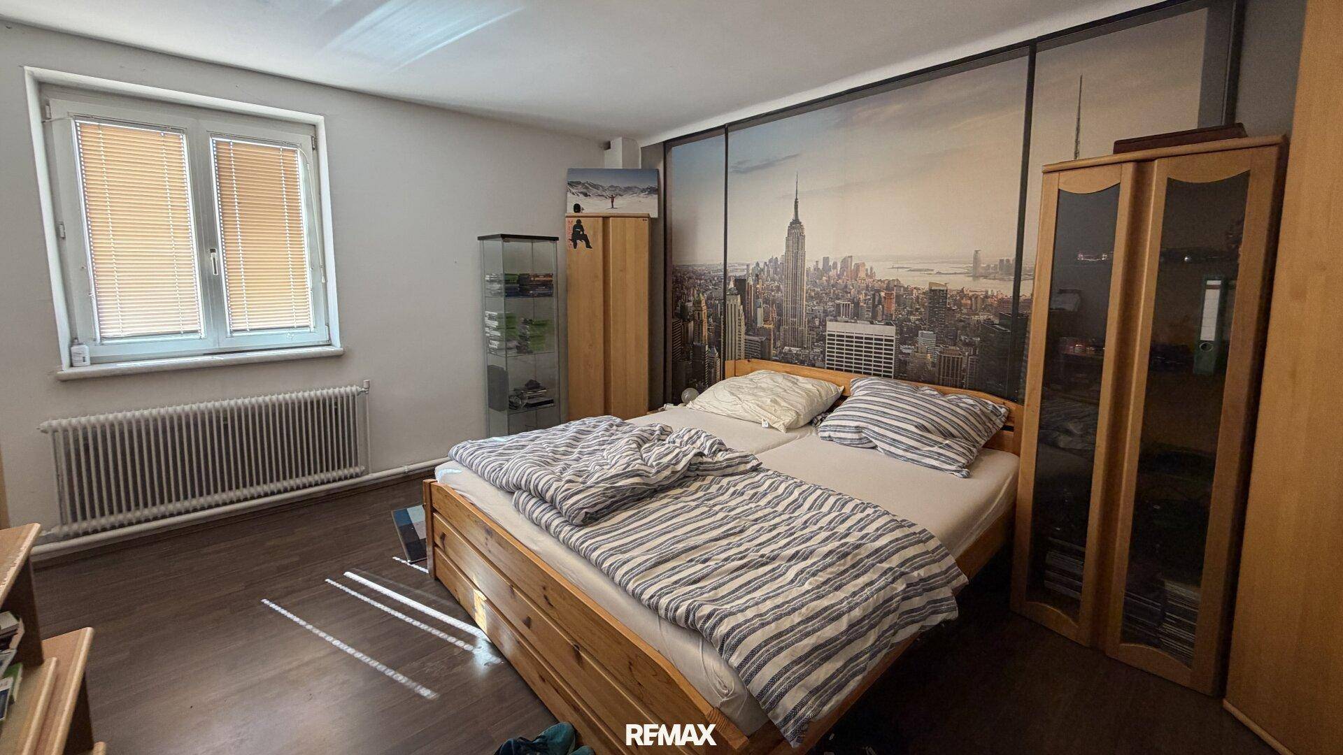 Schlafzimmer mit Doppelbett, Holzmöbeln und einer großen Wandtapete mit Stadtansicht.