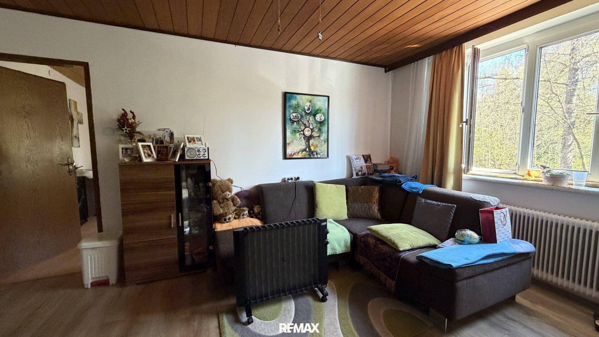 Gemütliches Wohnzimmer mit Ecksofa, Holzdecke und Fenster mit Blick ins Grüne.