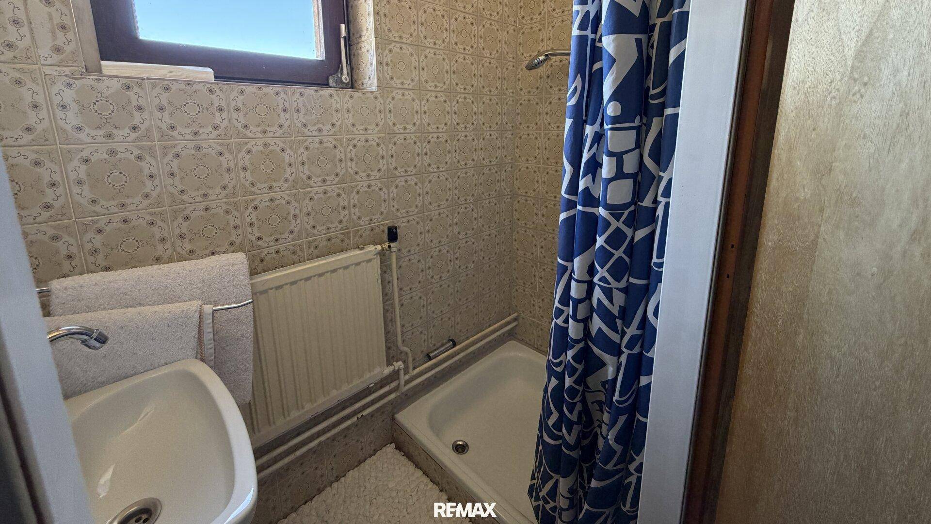 Kleines, gefliestes Badezimmer mit Dusche, Waschbecken und Fenster, in älterem Zustand.
