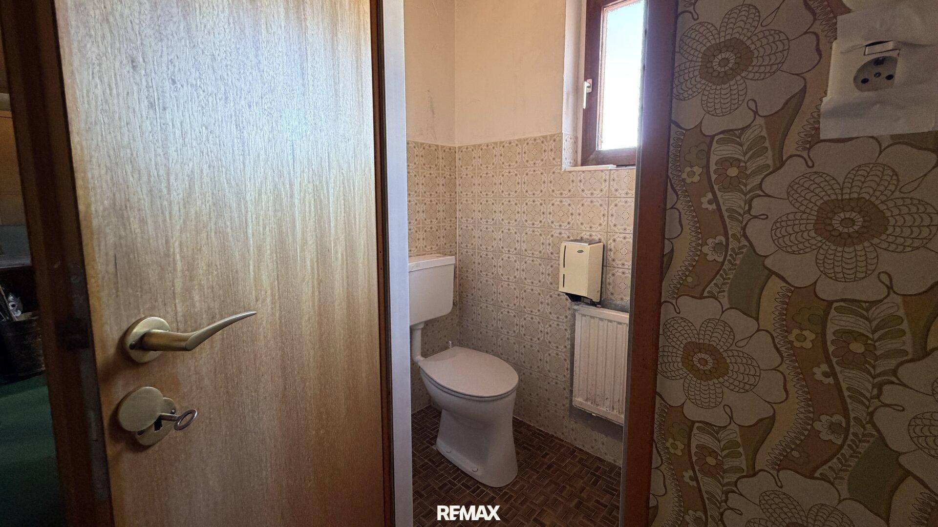 Separate Toilette mit Fenster und floraler Tapete, in einem älteren, aber funktionalen Zustand.