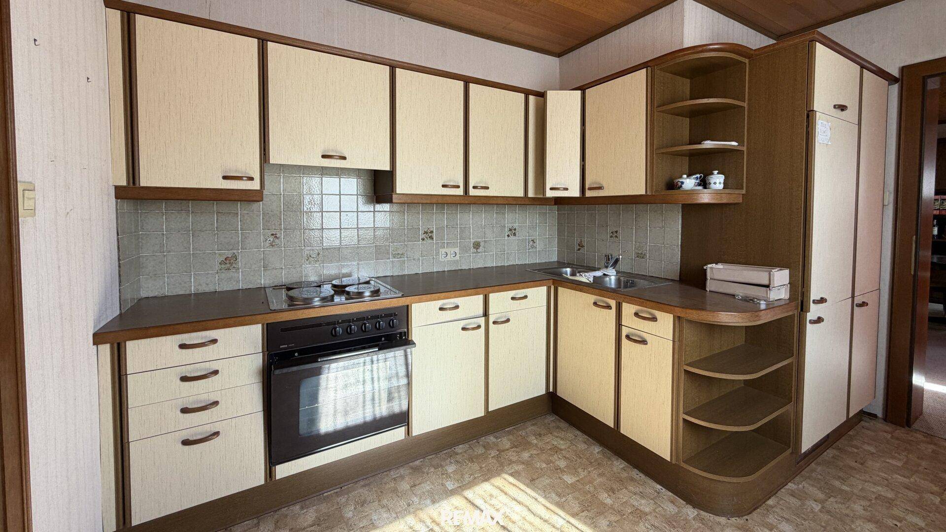 Ältere Küche mit beigefarbenen Schränken, Herd, Backofen und gefliestem Spritzschutz.