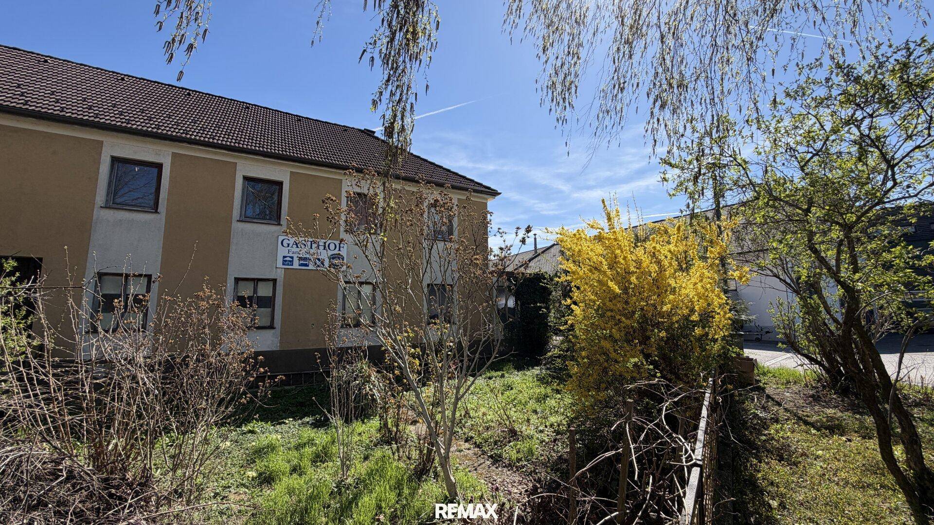 Gebäudeansicht eines Gasthofs mit Gartenbereich und blühenden Sträuchern unter blauem Himmel.