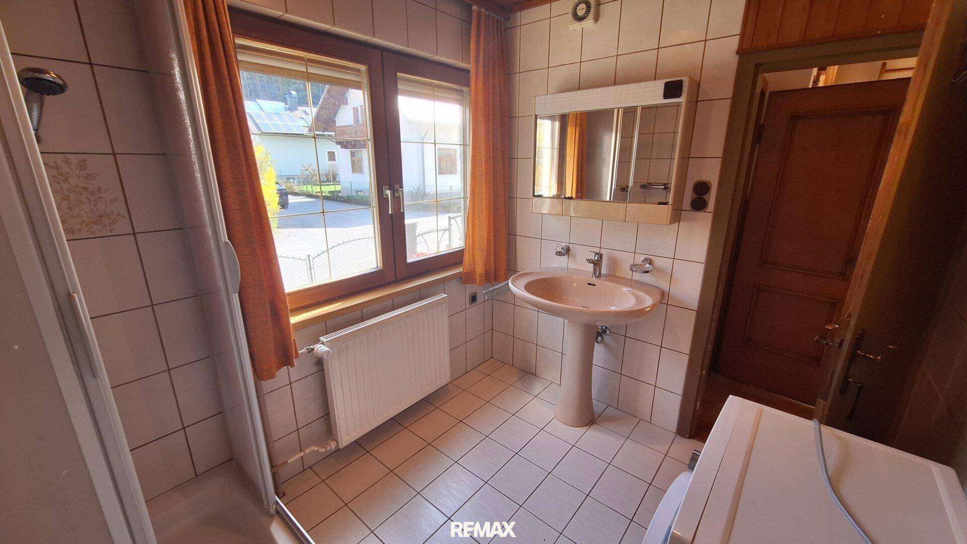 Helles Badezimmer mit weißen Fliesen, Duschkabine und Fenster mit Blick nach draußen.