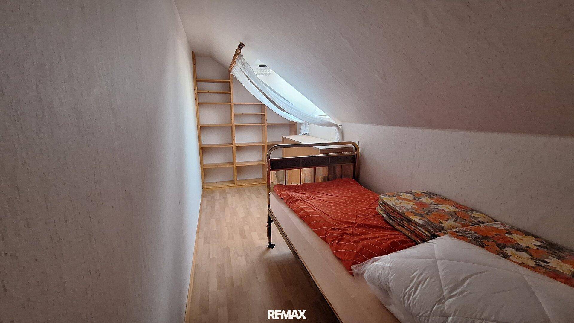 Kleines Schlafzimmer im Dachgeschoss mit Einzelbett, Holzregalen und schrägen Wänden.