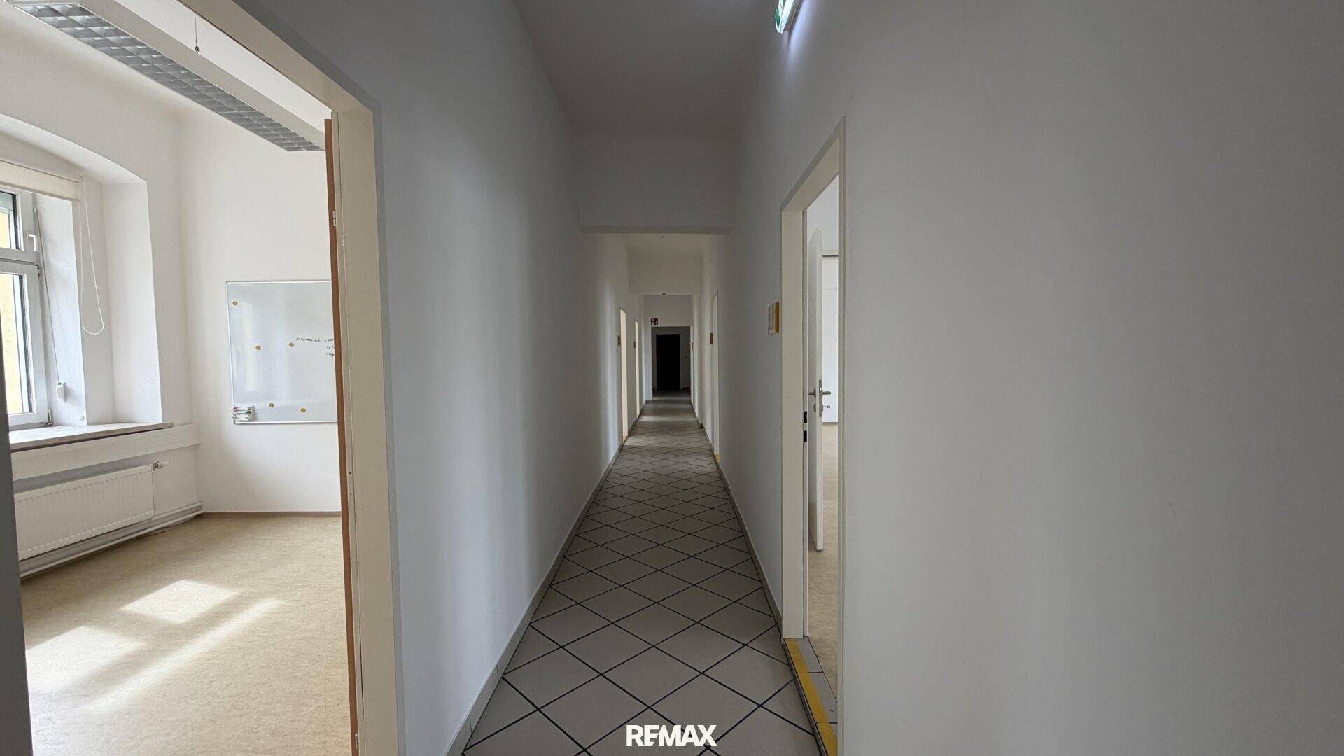 Heller Flur mit gefliestem Boden und Blick in einen angrenzenden Büroraum mit Fenstern.