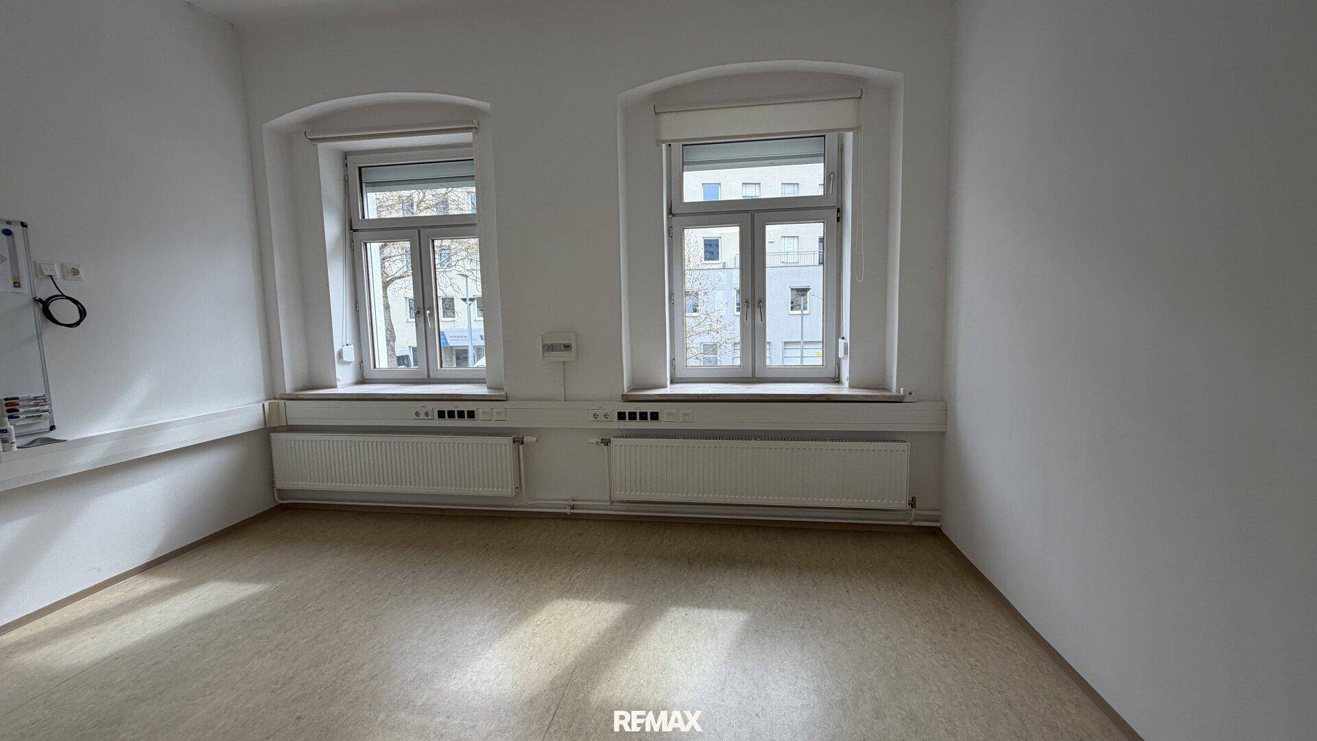 Leerer Büroraum mit zwei großen Fenstern, die für ausreichend Tageslicht und eine angenehme Arbeitsatmosphäre sorgen.