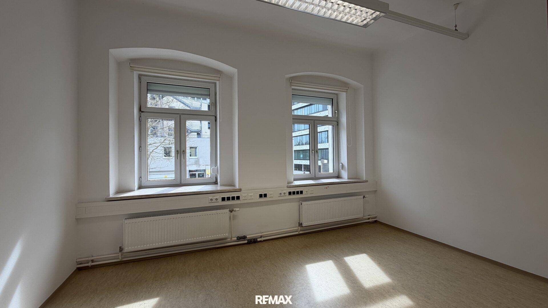 Leerer Büroraum mit zwei großen Fenstern, die viel Tageslicht hereinlassen und eine helle Atmosphäre schaffen.