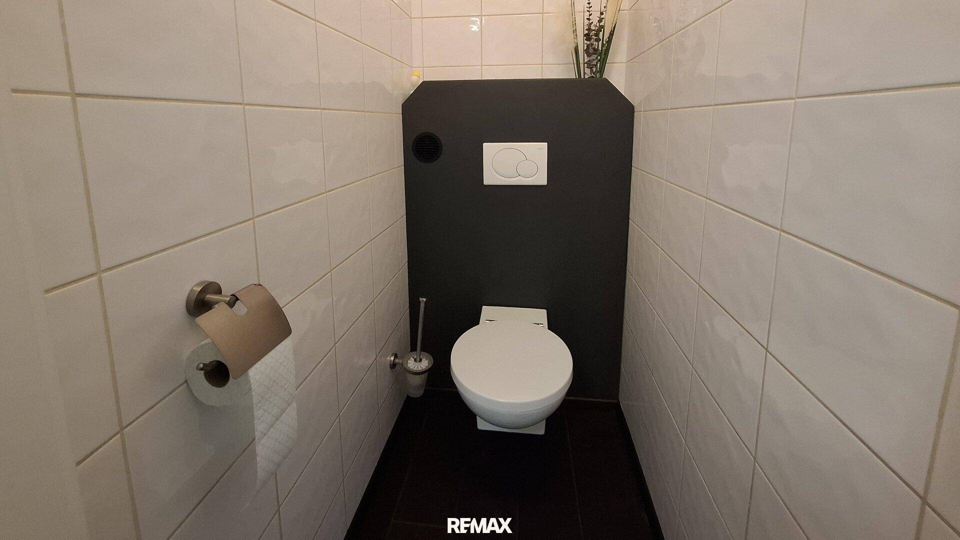 Separates WC mit weißen Fliesen, wandhängender Toilette und einer dunklen Akzentwand.