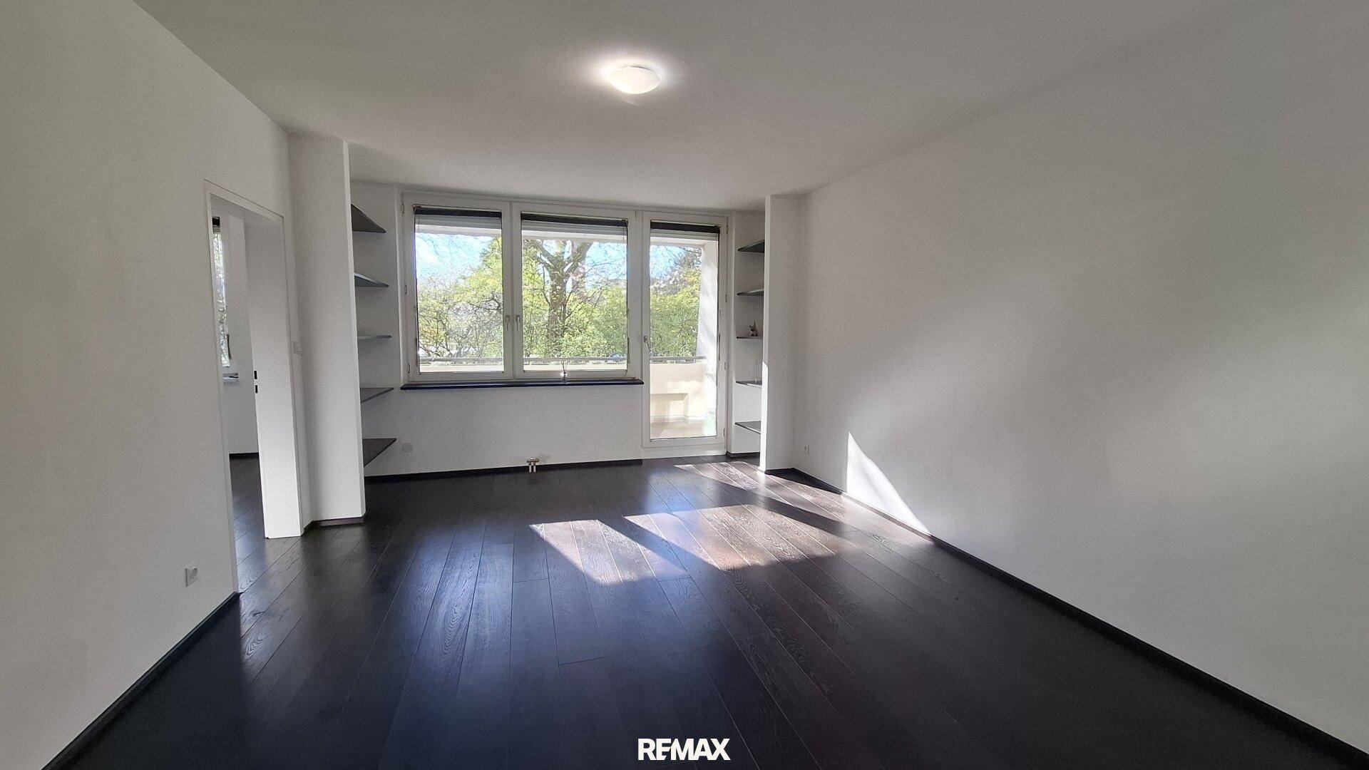 Helles, leeres Wohnzimmer mit dunklem Holzboden, weißen Wänden und großen Fenstern mit Blick ins Grüne.