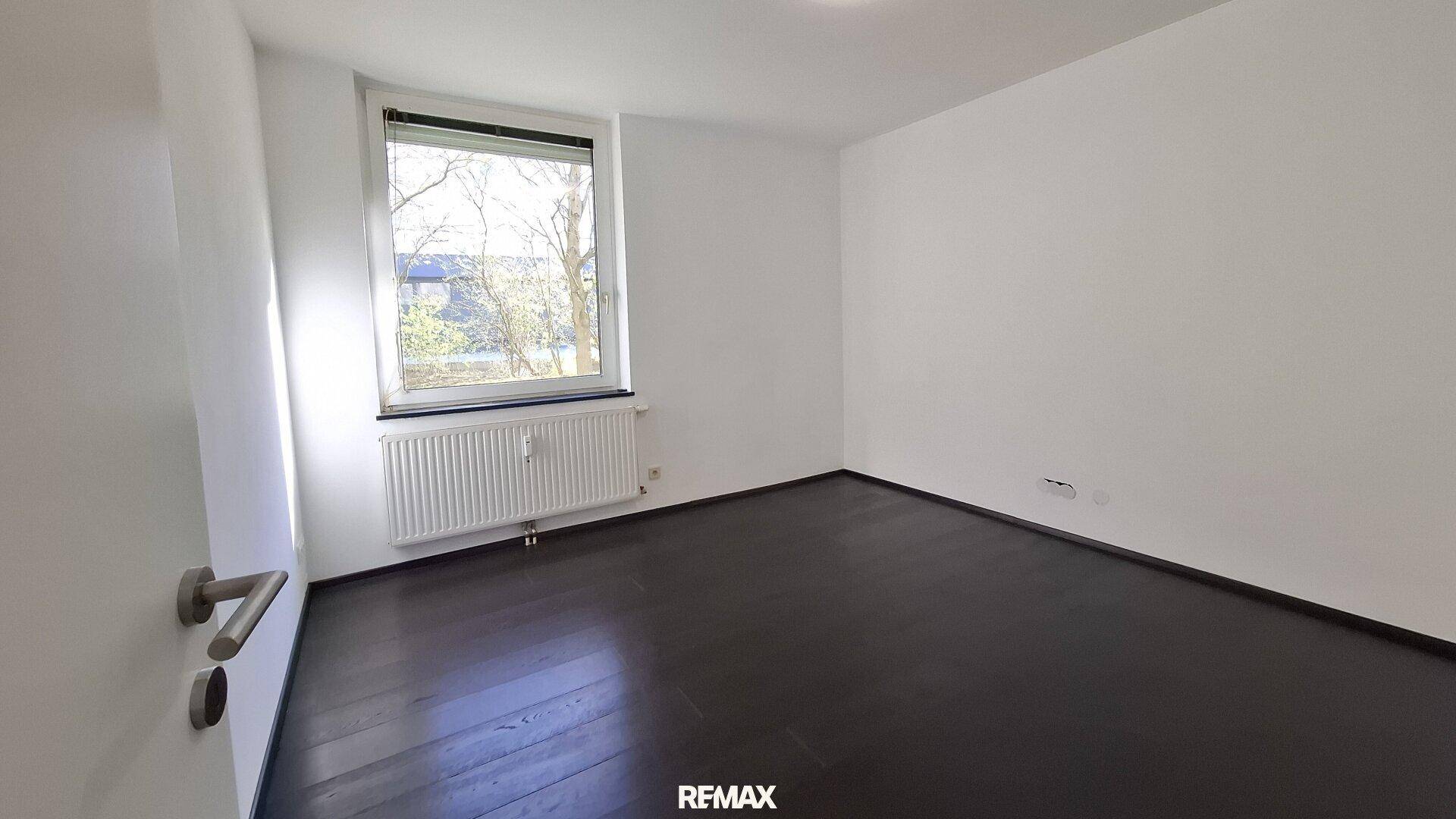 Helles, leeres Schlafzimmer mit dunklem Holzboden, weißen Wänden und einem Fenster mit Heizkörper.