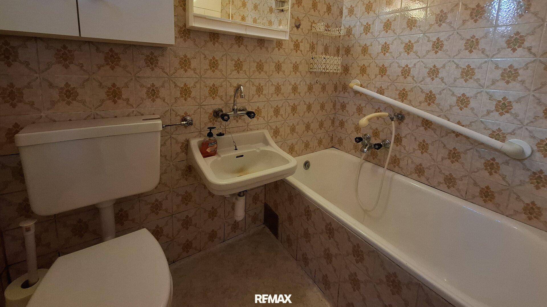 Badezimmer mit Badewanne, WC, Waschbecken und Haltegriffen, ausgestattet mit gemusterten Fliesen.