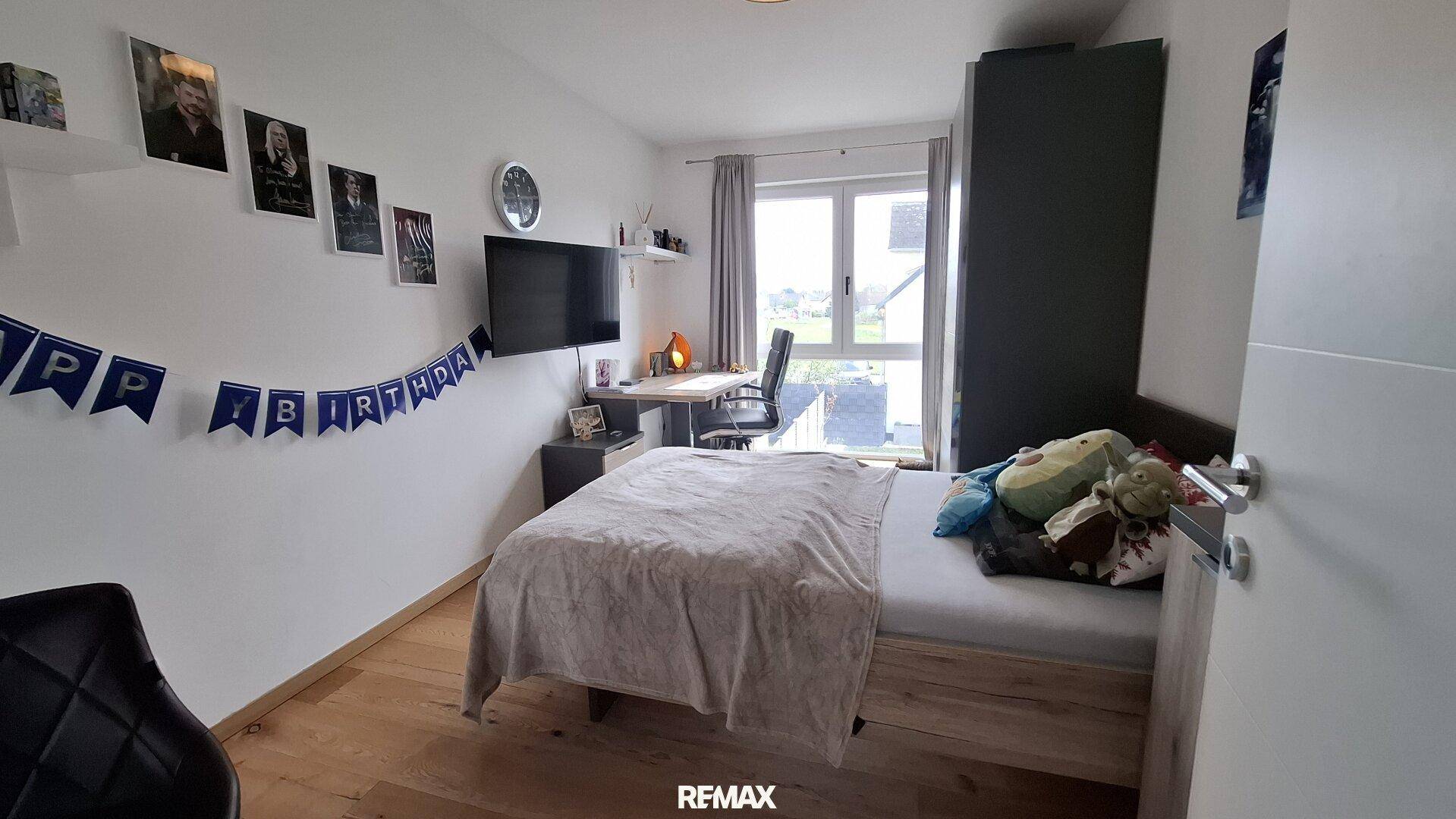 Helles Schlafzimmer mit Einzelbett, Schreibtisch am Fenster und Wandfernseher.