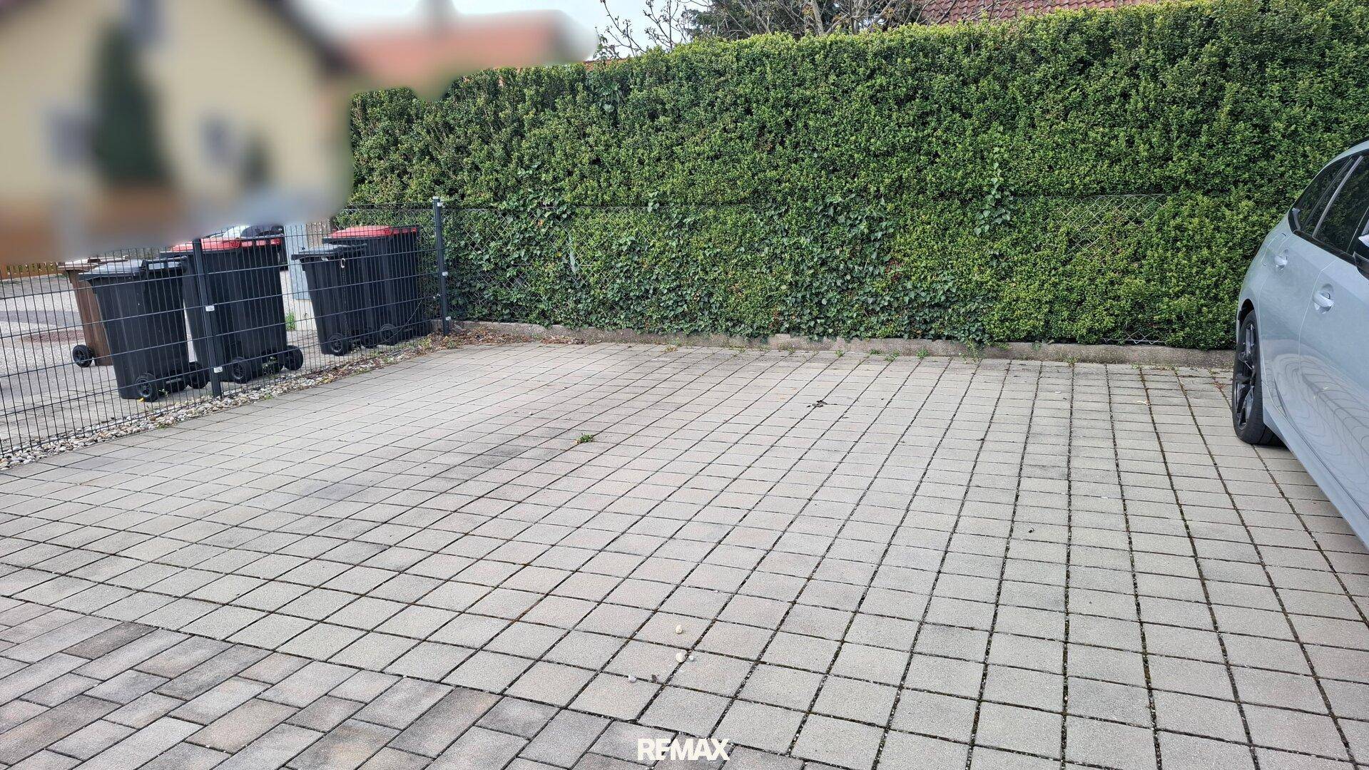Gepflasterter Außenparkplatz neben einer Hecke mit Mülltonnen.