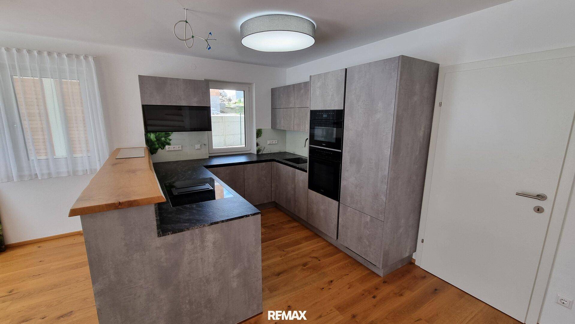 Moderne Küche mit grauen Schränken, dunklen Arbeitsplatten, Holzboden und integrierter Frühstücksbar.