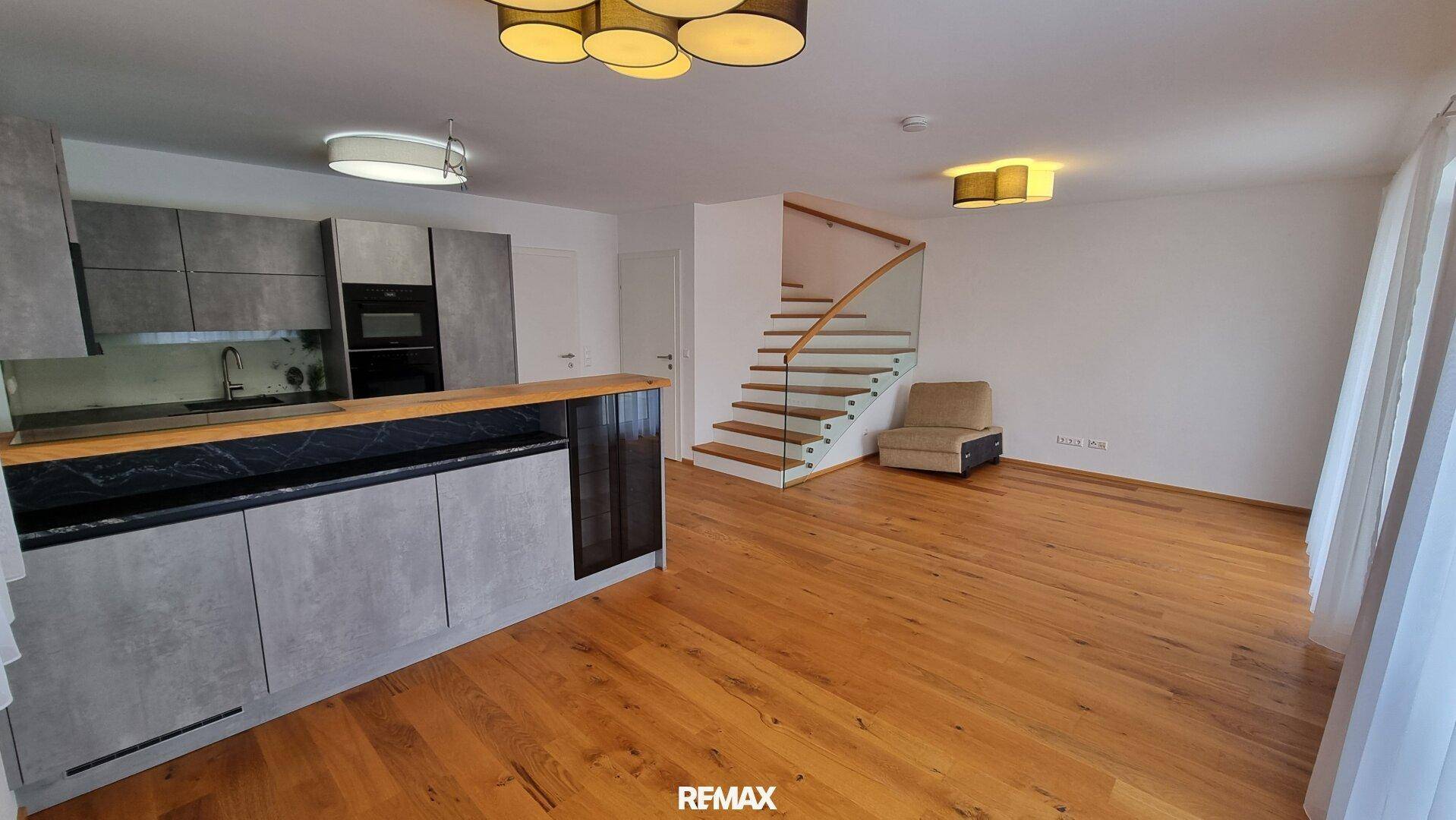 Offener Wohnbereich mit integrierter Küche, Holzboden und moderner Treppe mit Glasgeländer.
