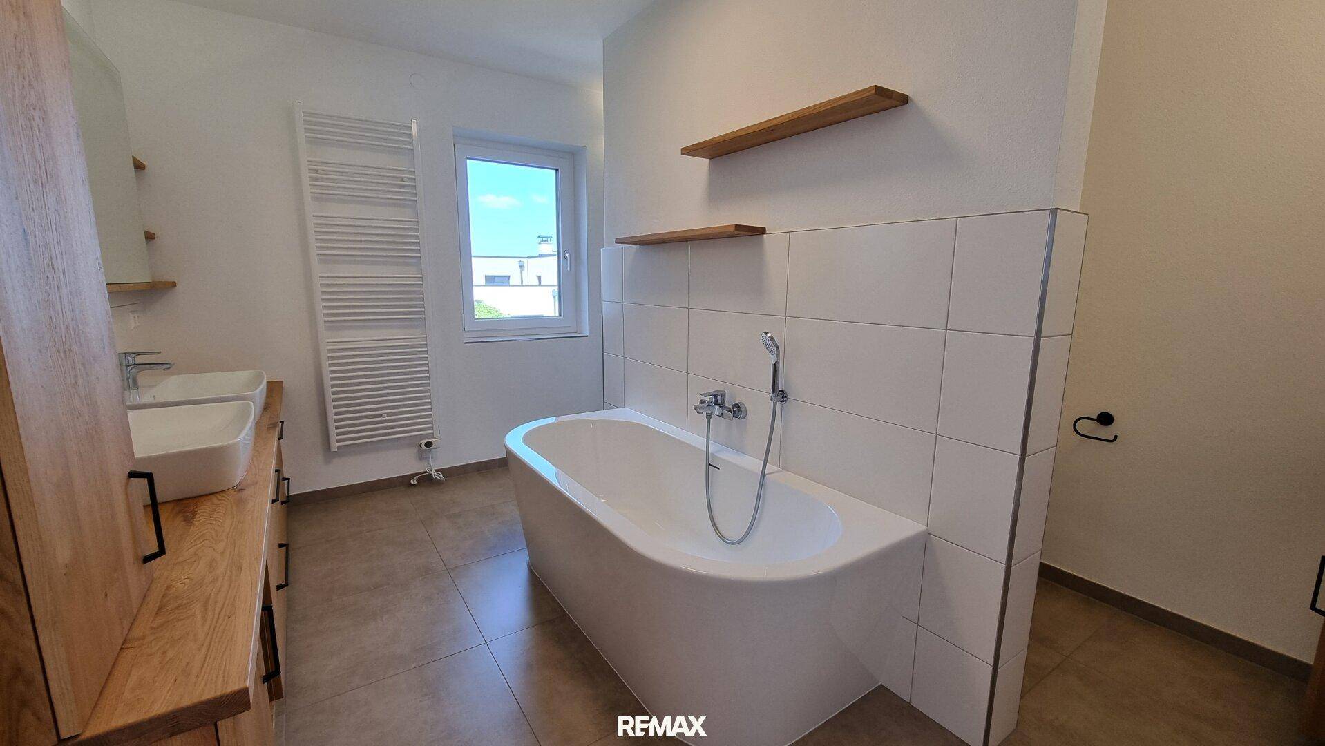 Helles Badezimmer mit freistehender Badewanne, Doppelwaschbecken, beheiztem Handtuchhalter und Fenster.