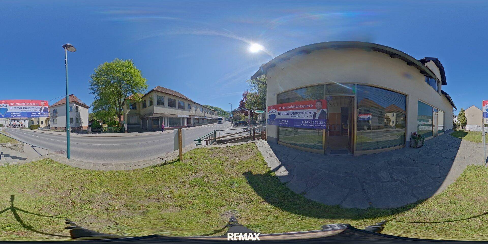 Panoramablick auf das Gebäude von der Straße aus, mit umliegenden Häusern und viel Grün.