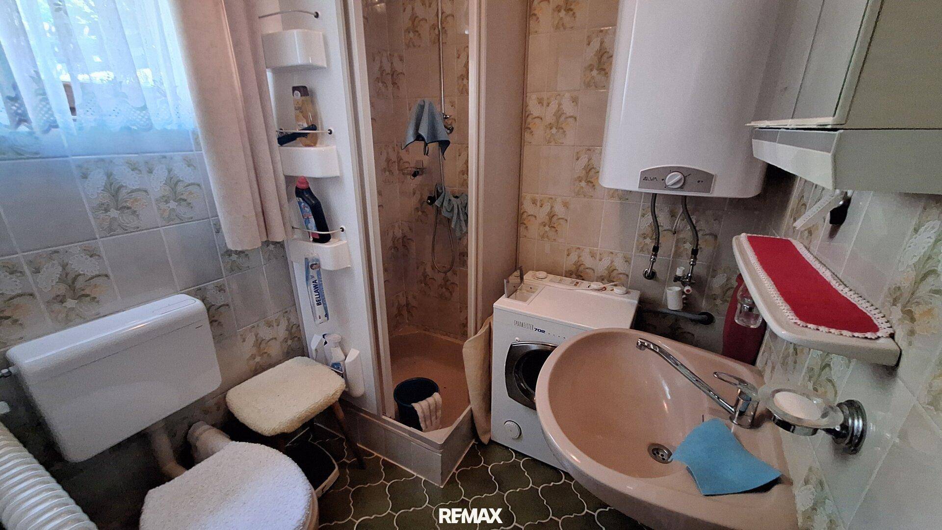 Kleines Badezimmer mit Dusche, WC und Waschmaschine, ausgestattet mit beigen Fliesen.