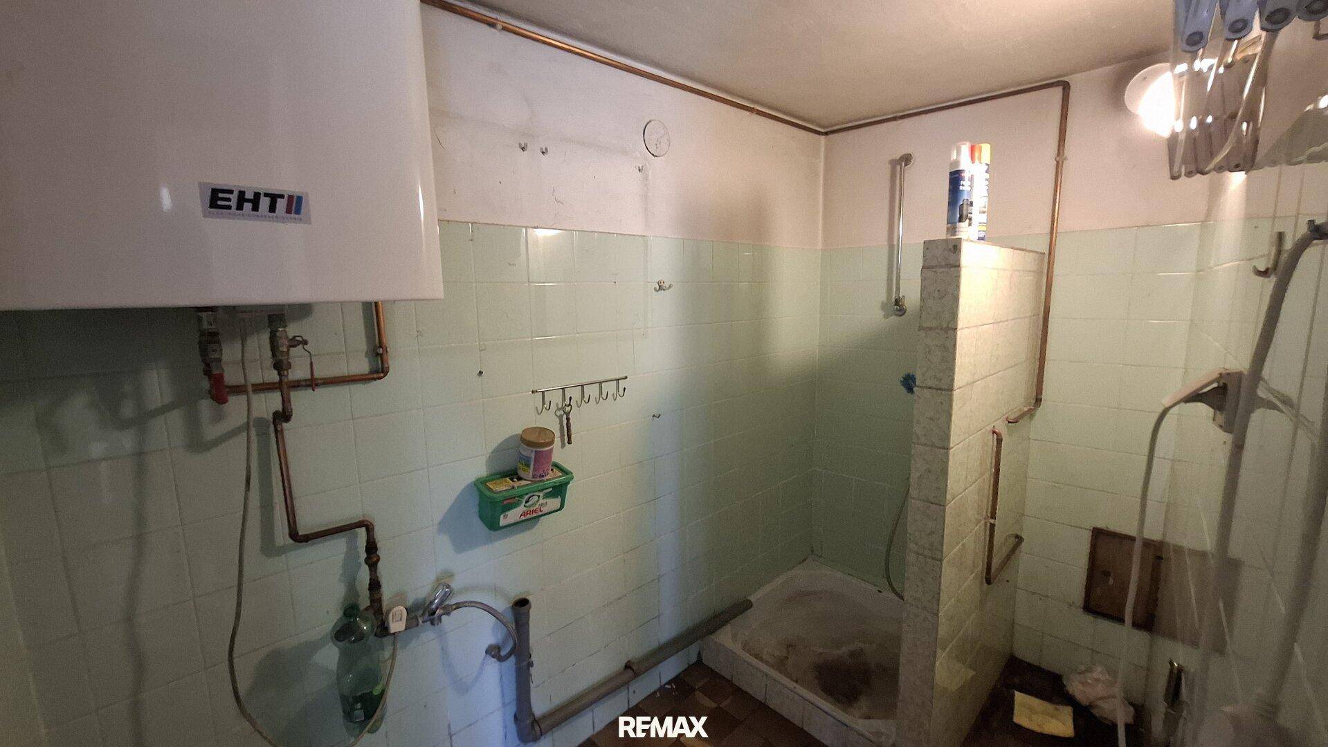 Altes Badezimmer mit hellgrünen Fliesen, Duschwanne und einem Warmwasserboiler an der Wand.