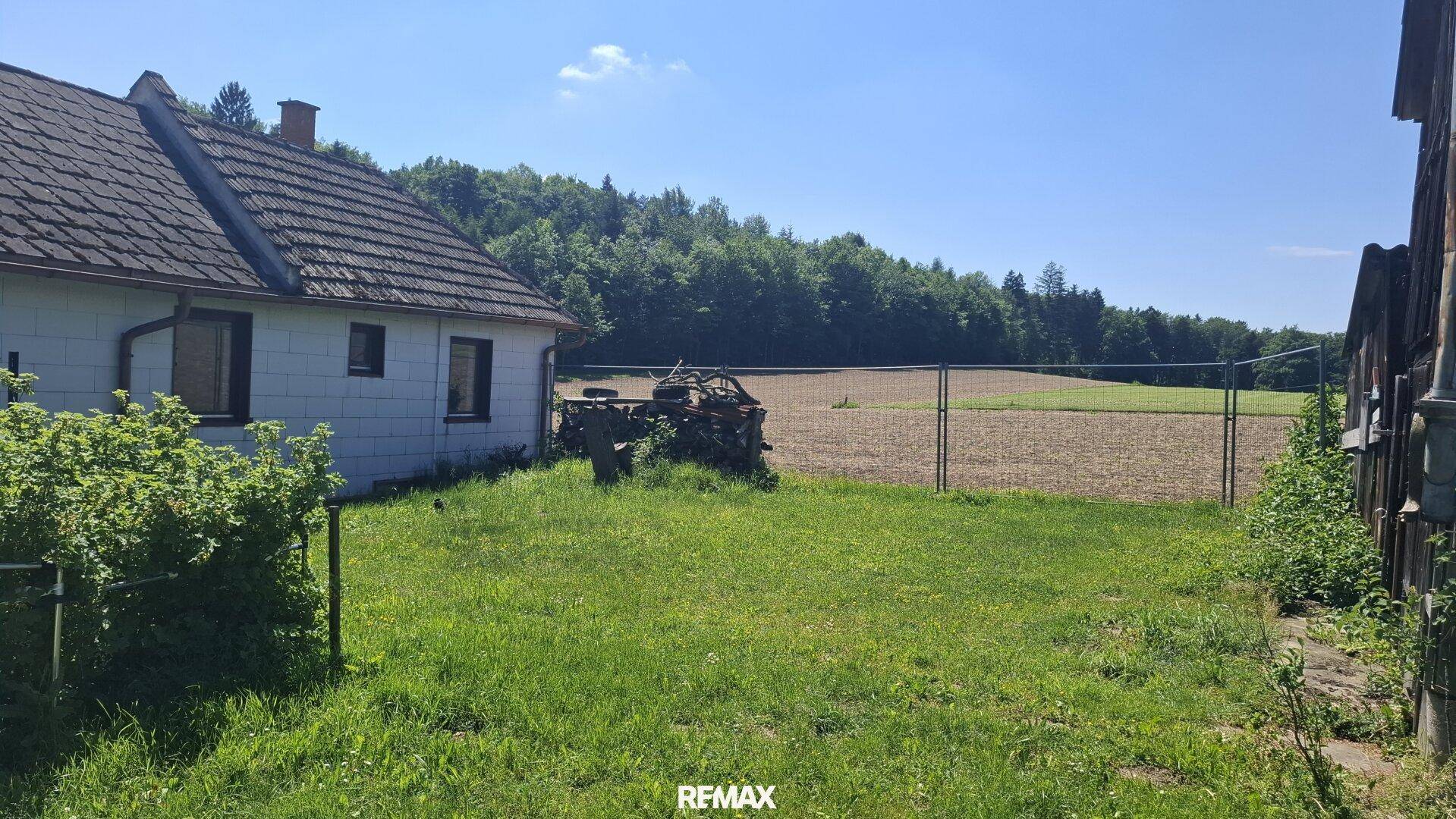 Ländliches Haus mit Ziegeldach, umgeben von einer Wiese und weiten Feldern unter blauem Himmel.