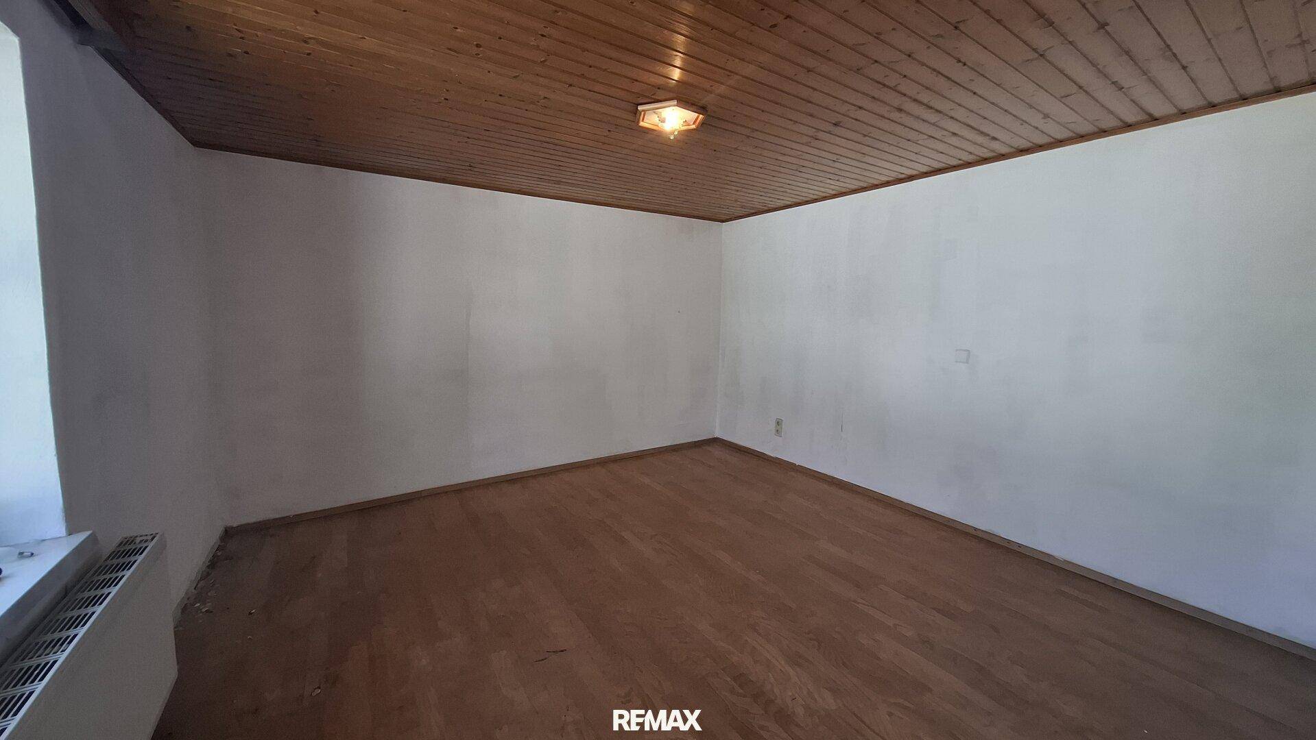 Leerer Raum mit Holzdielenboden, Holzdecke und einem Heizkörper unter dem Fenster.