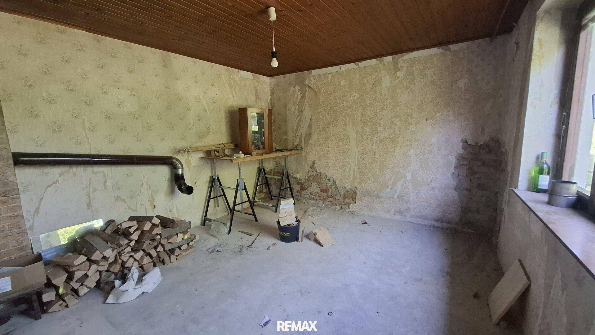 Raum in schlechtem Zustand mit abblätternder Tapete, Betonboden und einem Stapel Brennholz.