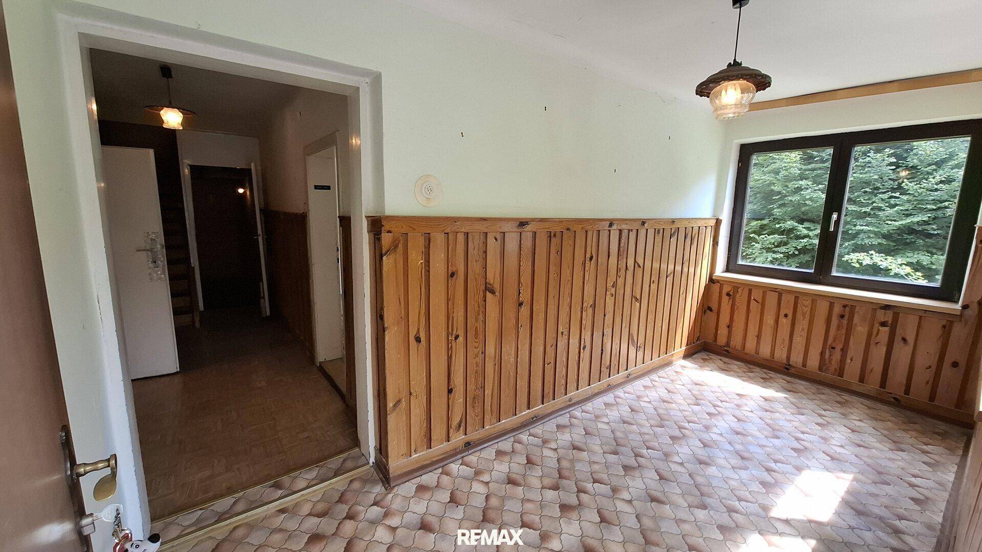 Zimmer mit Holzvertäfelung, gemustertem Boden und Fenster mit Blick ins Grüne.