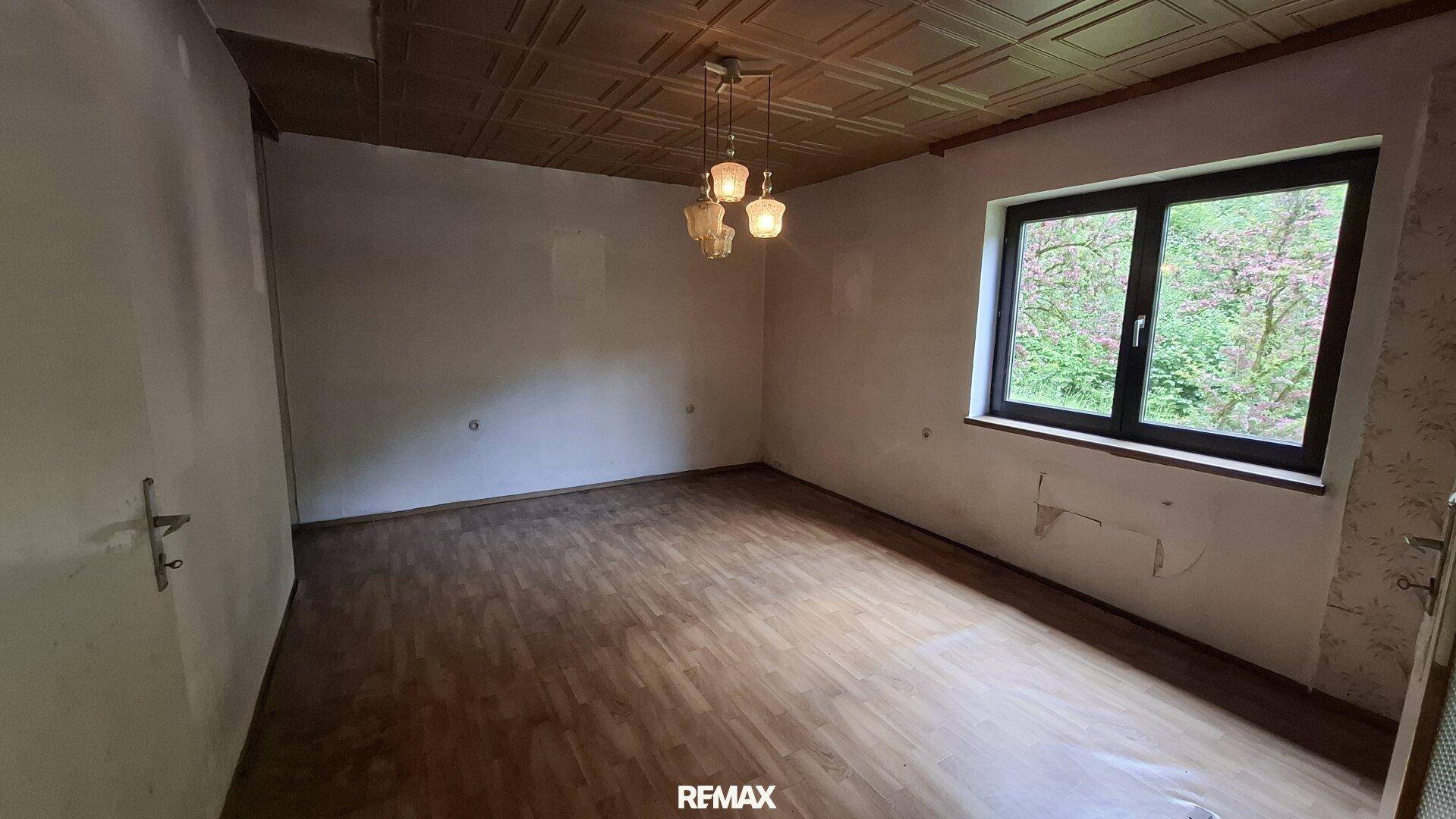 Leeres Zimmer mit Holzdecke, Holzboden und Fenster mit Blick auf Bäume.