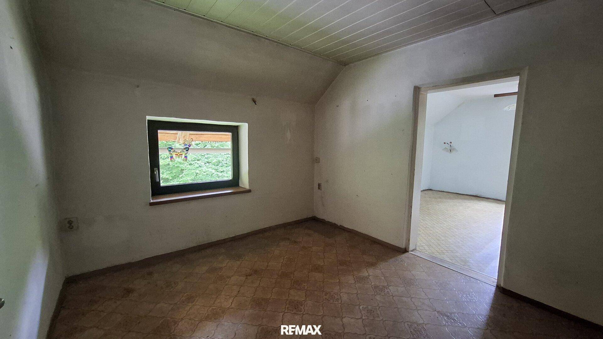 Kleines Zimmer mit Dachschräge, gemustertem Boden und Fenster mit Blick ins Freie.