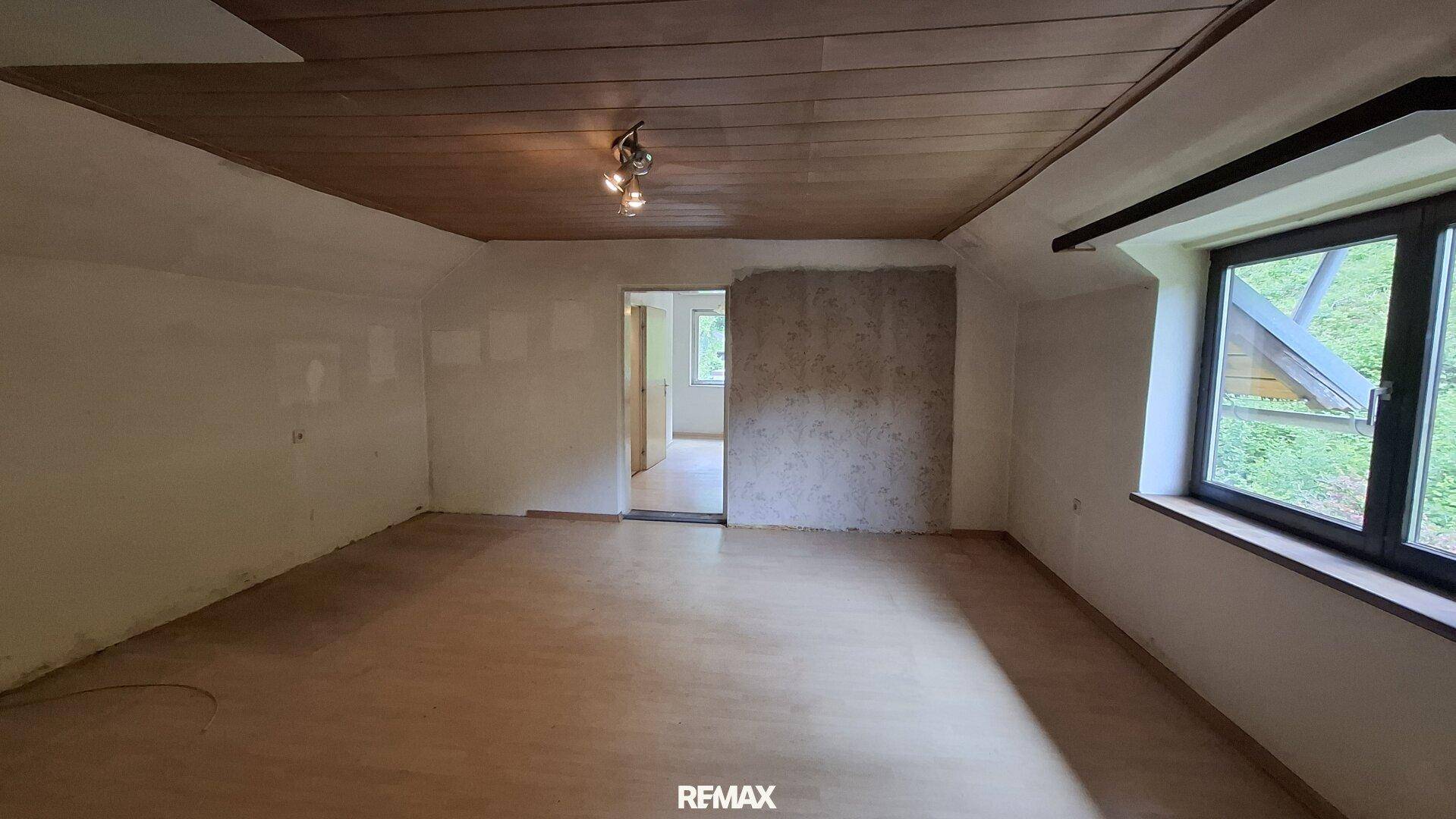 Geräumiges Zimmer mit Holzpaneeldecke, Holzboden und Fenster mit Blick ins Grüne.