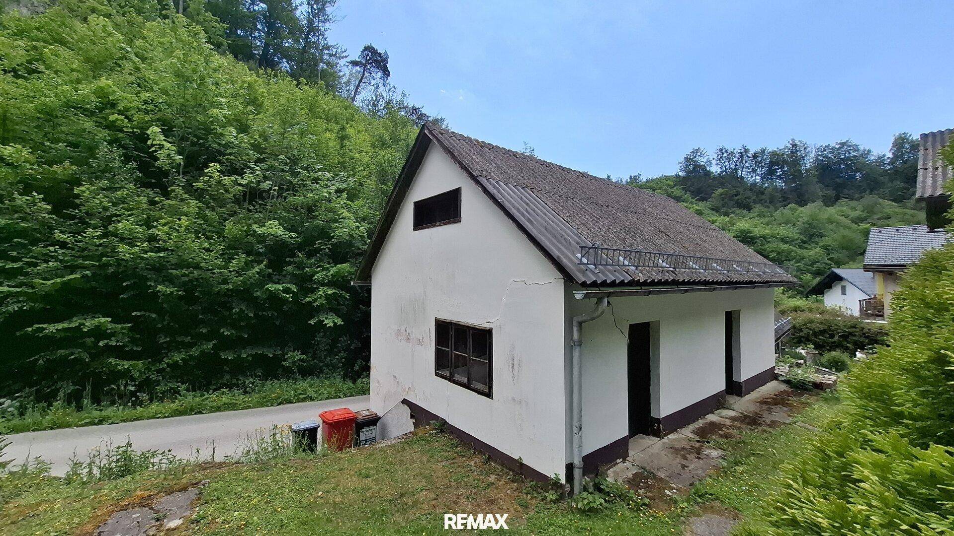 Seitenansicht eines einfachen Gebäudes mit Satteldach und Fenstern, umgeben von Grün.