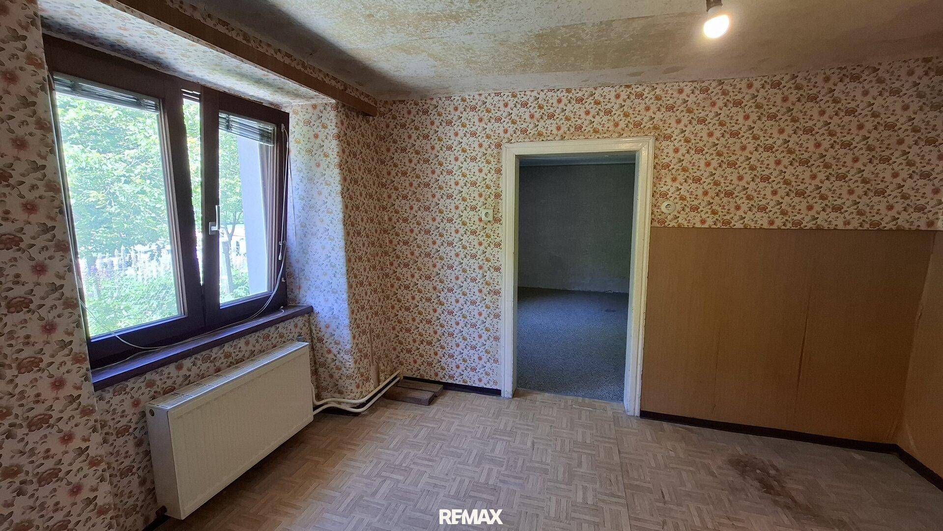 Zimmer mit gemusterter Tapete, Parkettboden und einem Heizkörper unter dem Fenster.