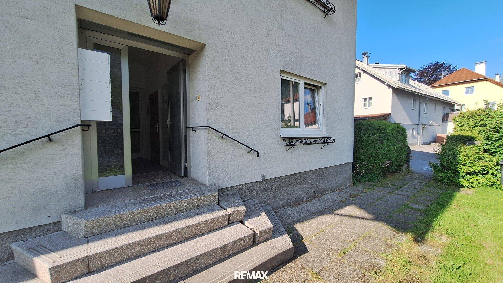 Eingangsbereich des Gebäudes mit Steinstufen, Handlauf und einer grünen Hecke.