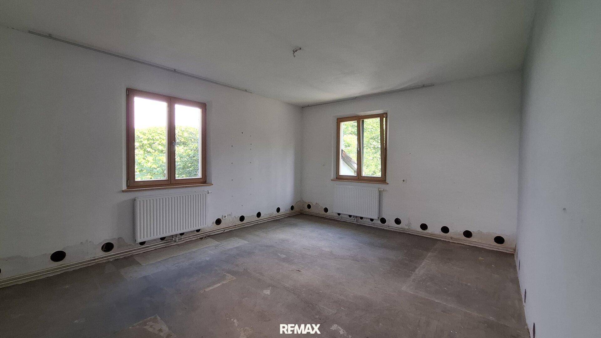Helles Zimmer mit zwei Fenstern, Heizkörpern und Blick in die Natur.