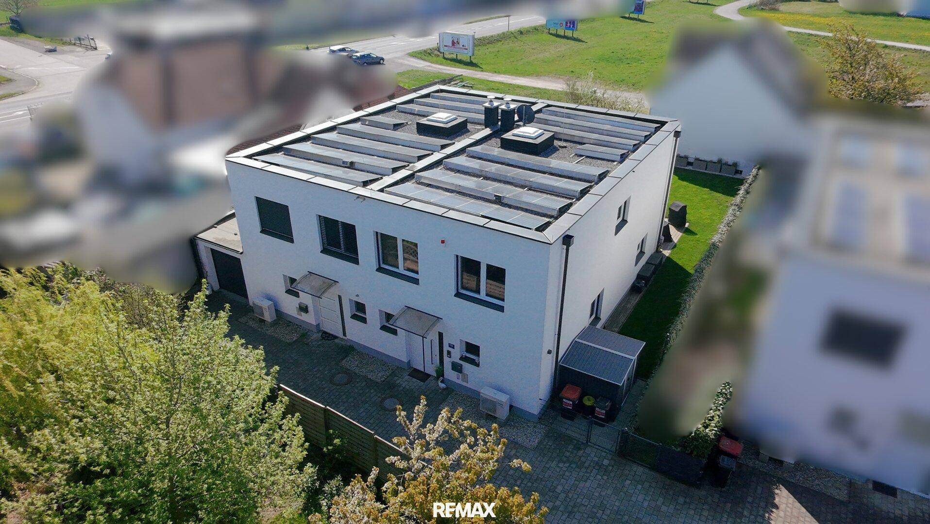 Modernes weißes Gebäude mit Flachdach und Solaranlagen, umgeben von einem kleinen Garten.