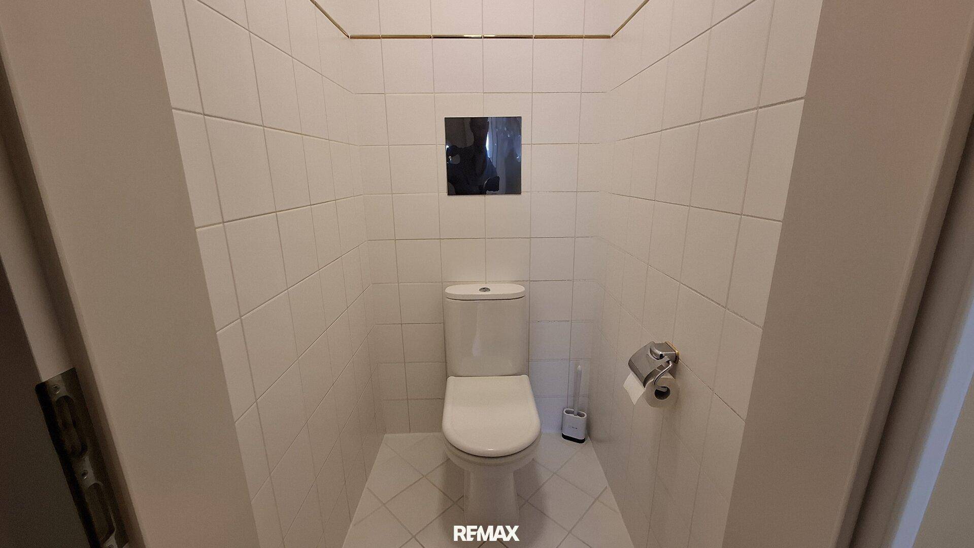 Kleines separates WC mit weiß gefliesten Wänden und Boden, ausgestattet mit einer Wandtoilette.