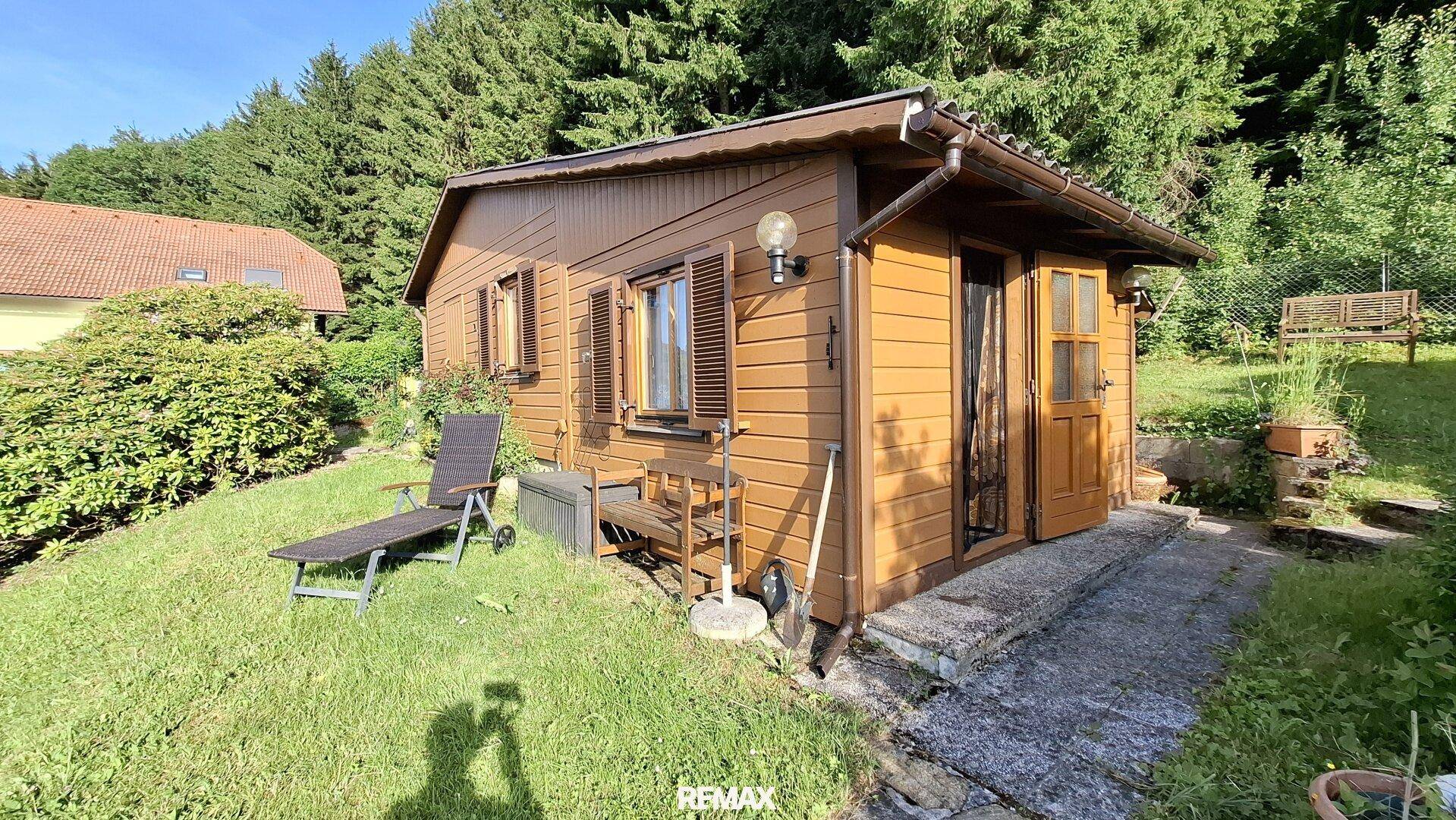 Holzhaus im Grünen mit kleiner Terrasse und Liegestuhl, umgeben von Bäumen.