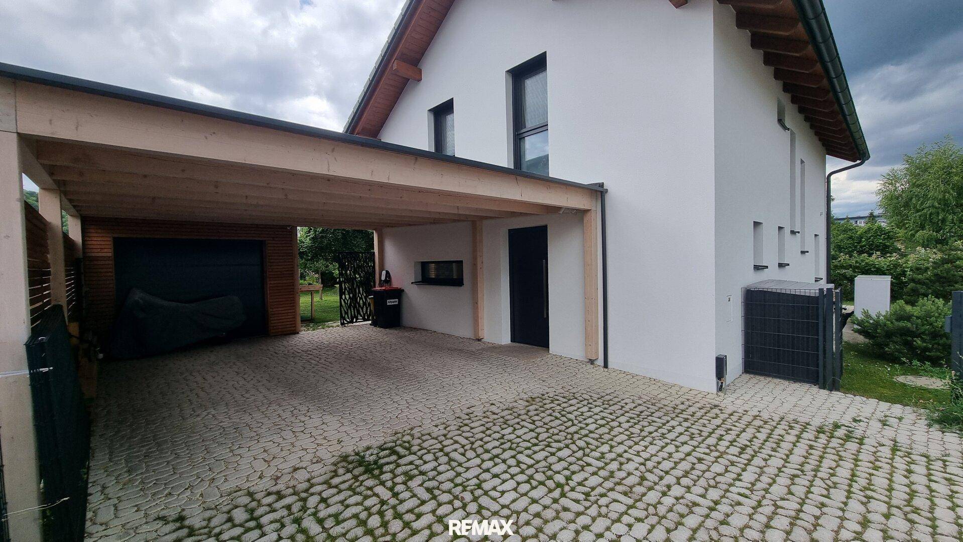Seitenansicht des Hauses mit Holz-Carport, gepflastertem Vorplatz und Eingangsbereich.