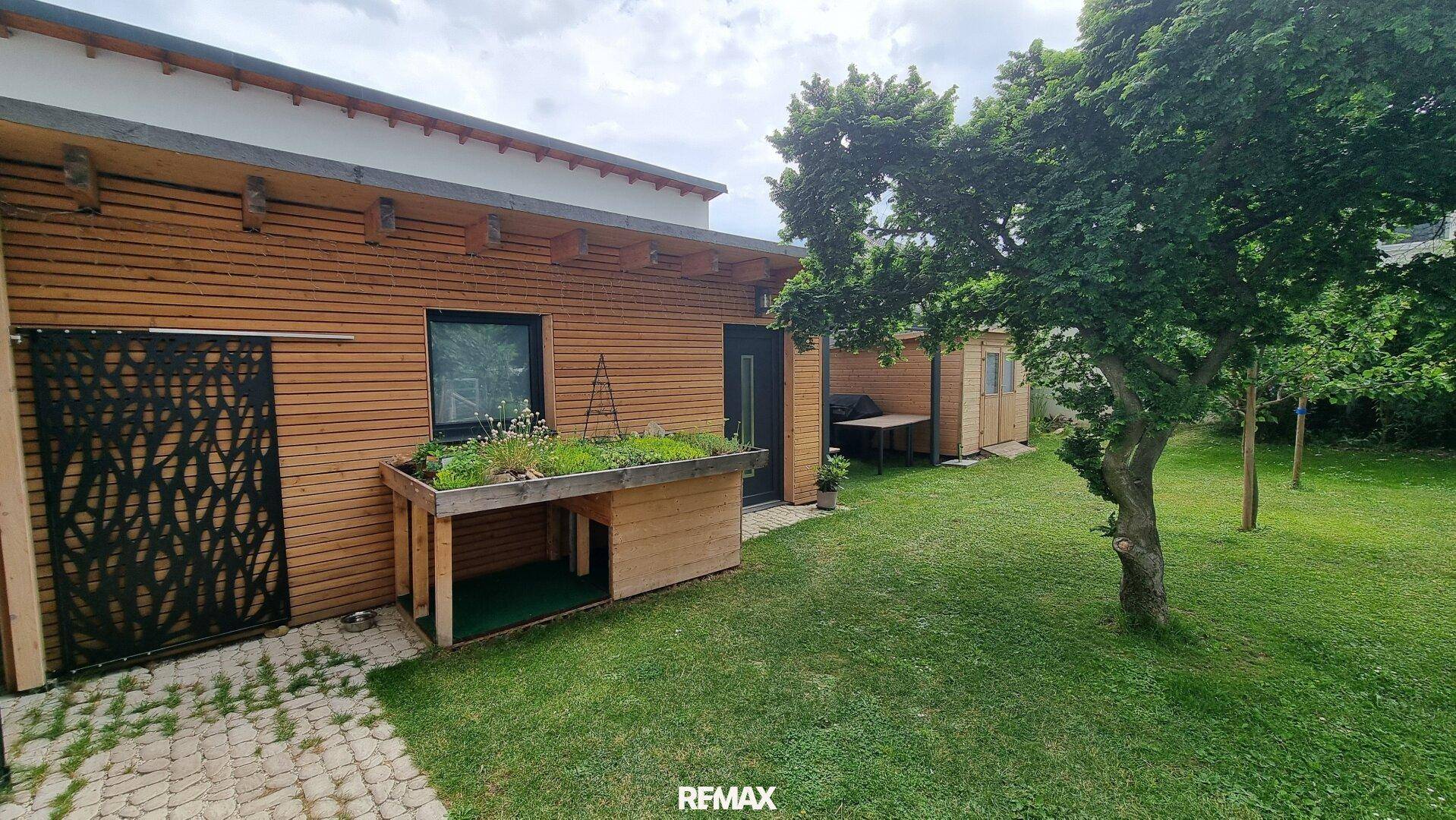 Seitenansicht des Hauses mit Holzfassade, kleinem Hochbeet und gepflegter Rasenfläche.