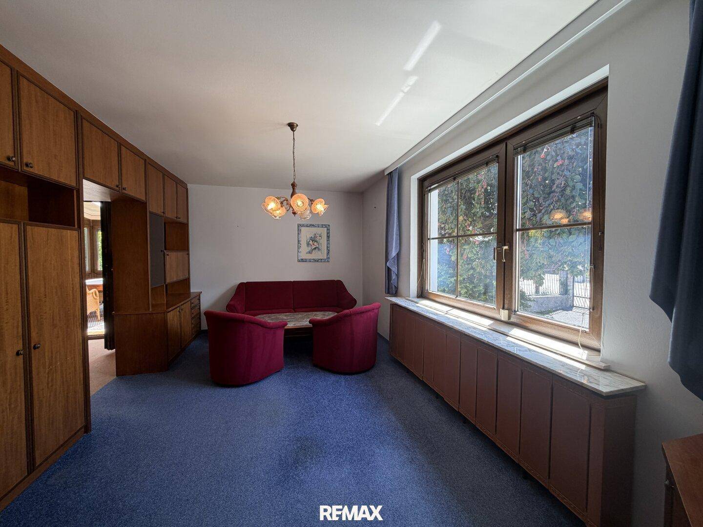 Geräumiges Wohnzimmer mit blauem Teppichboden, Holzschrankwand und großen Fenstern.