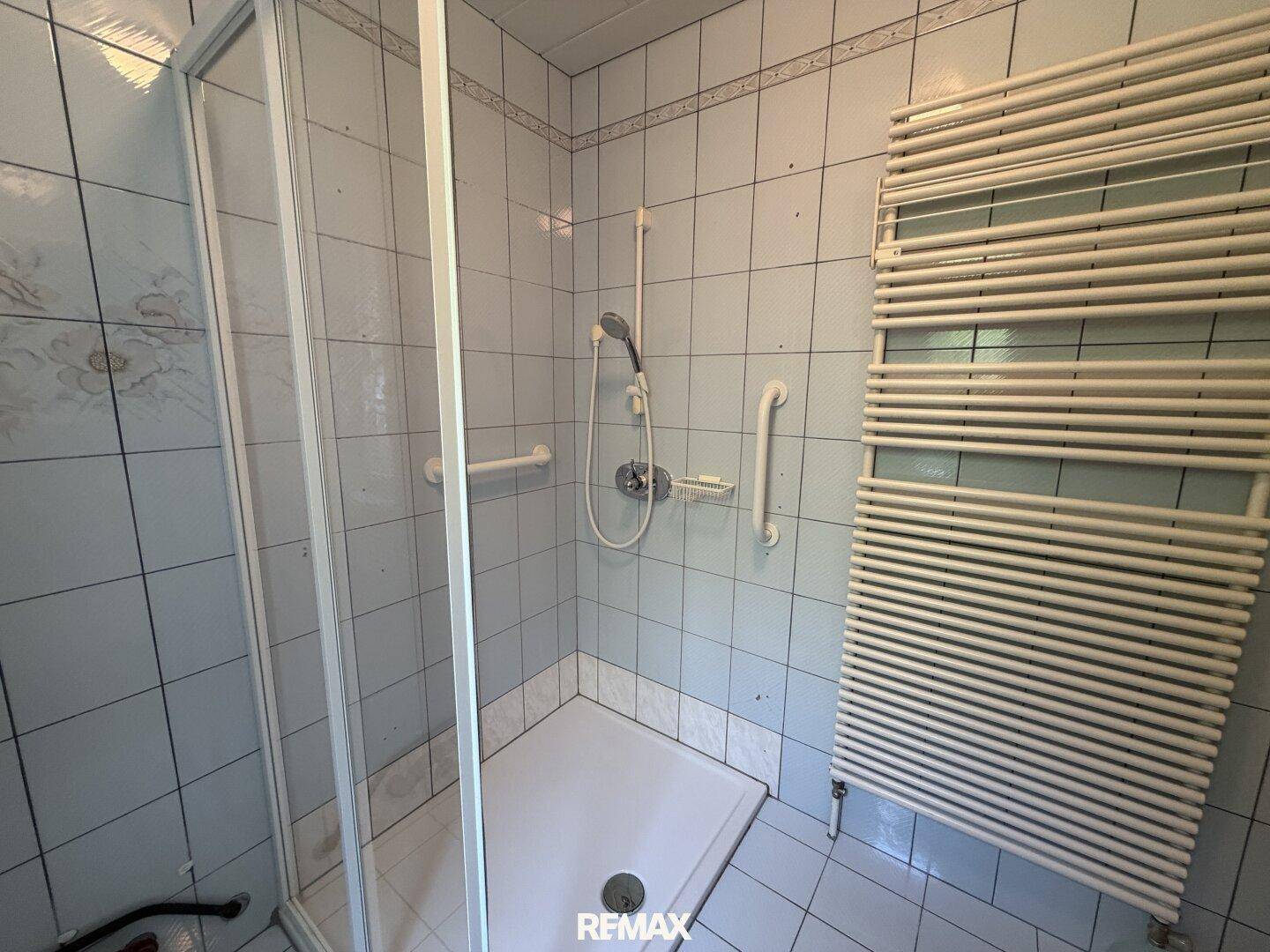 Duschbereich im Badezimmer mit Fliesenwänden, Duschkabine und Haltegriffen.