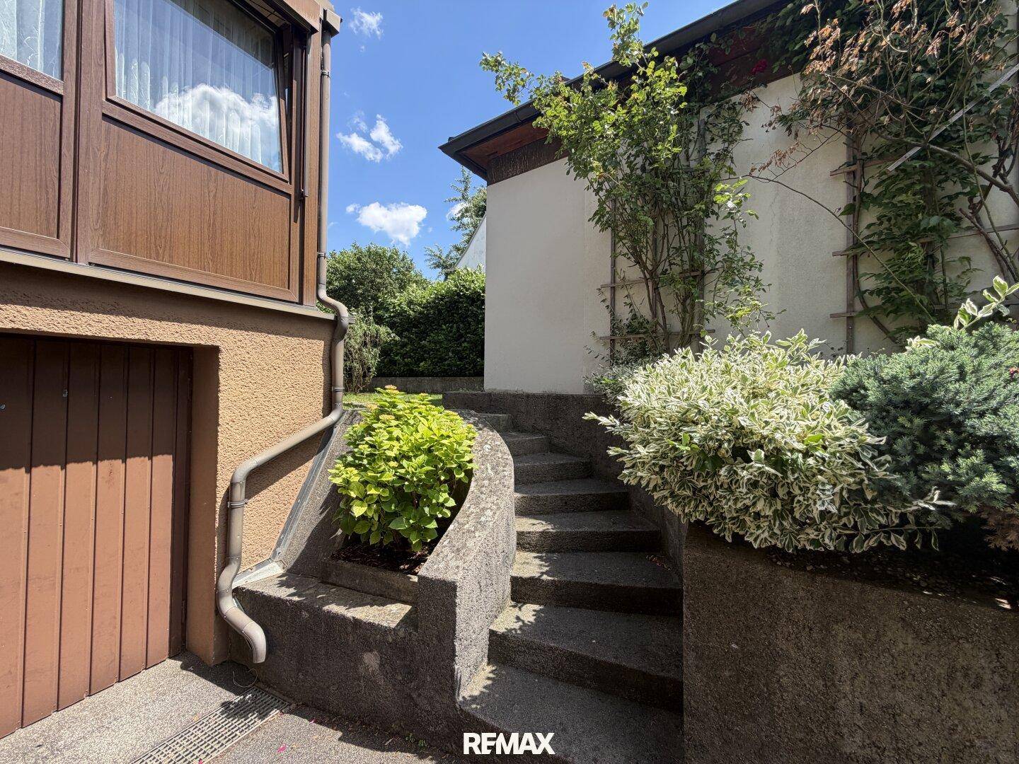 Außentreppe aus Beton, umgeben von Pflanzen, führt zum Eingangsbereich des Hauses neben der Garage.