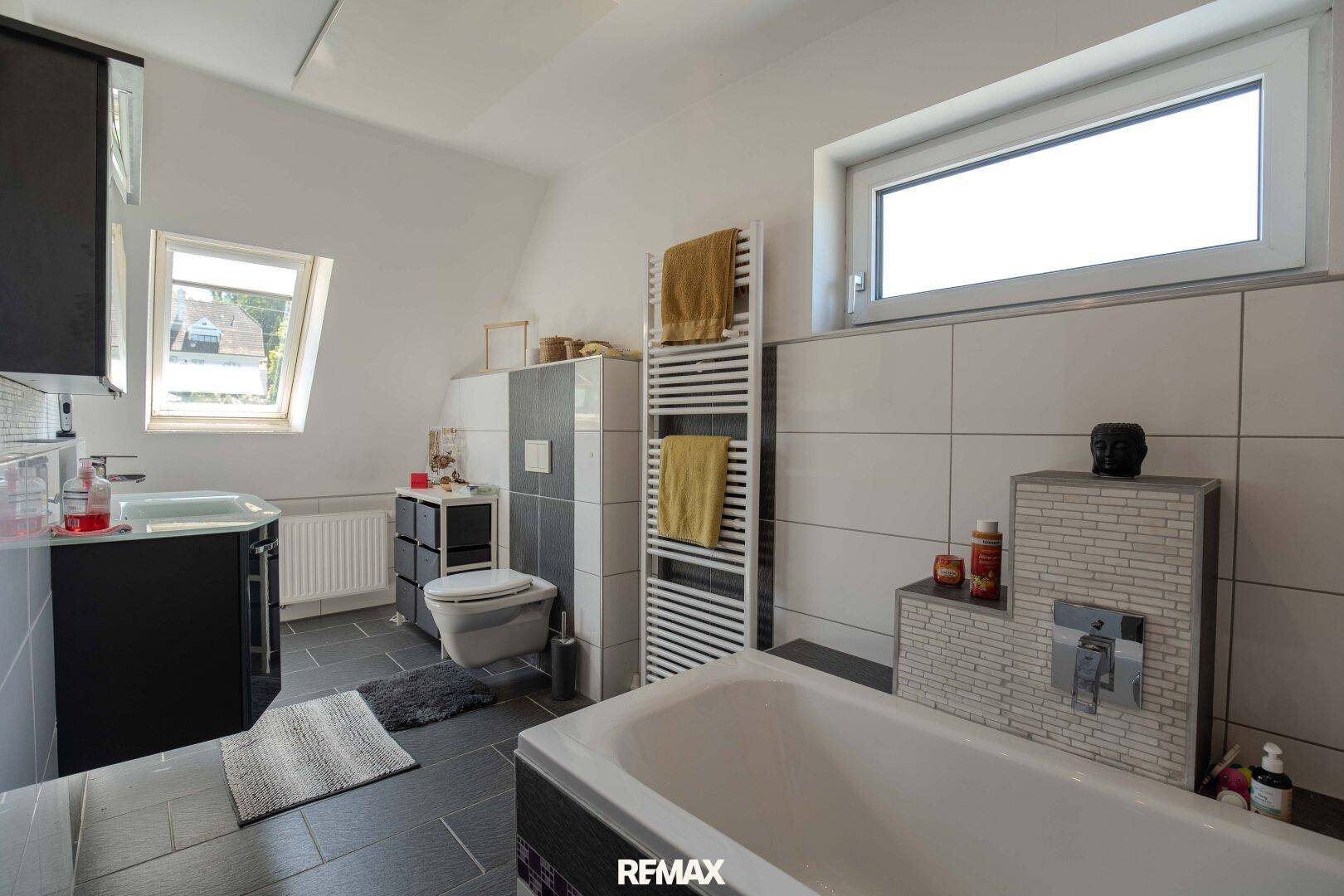 Modernes Badezimmer mit Badewanne, WC, Waschbecken und zwei Fenstern.