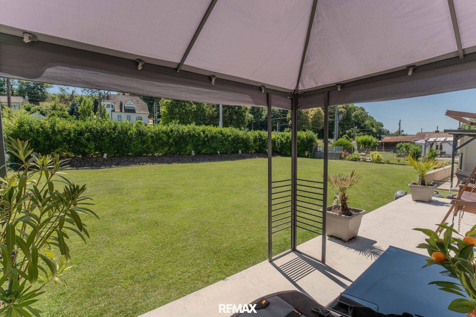 Gepflegter Garten mit Rasenfläche, Hecke und überdachtem Sitzbereich.