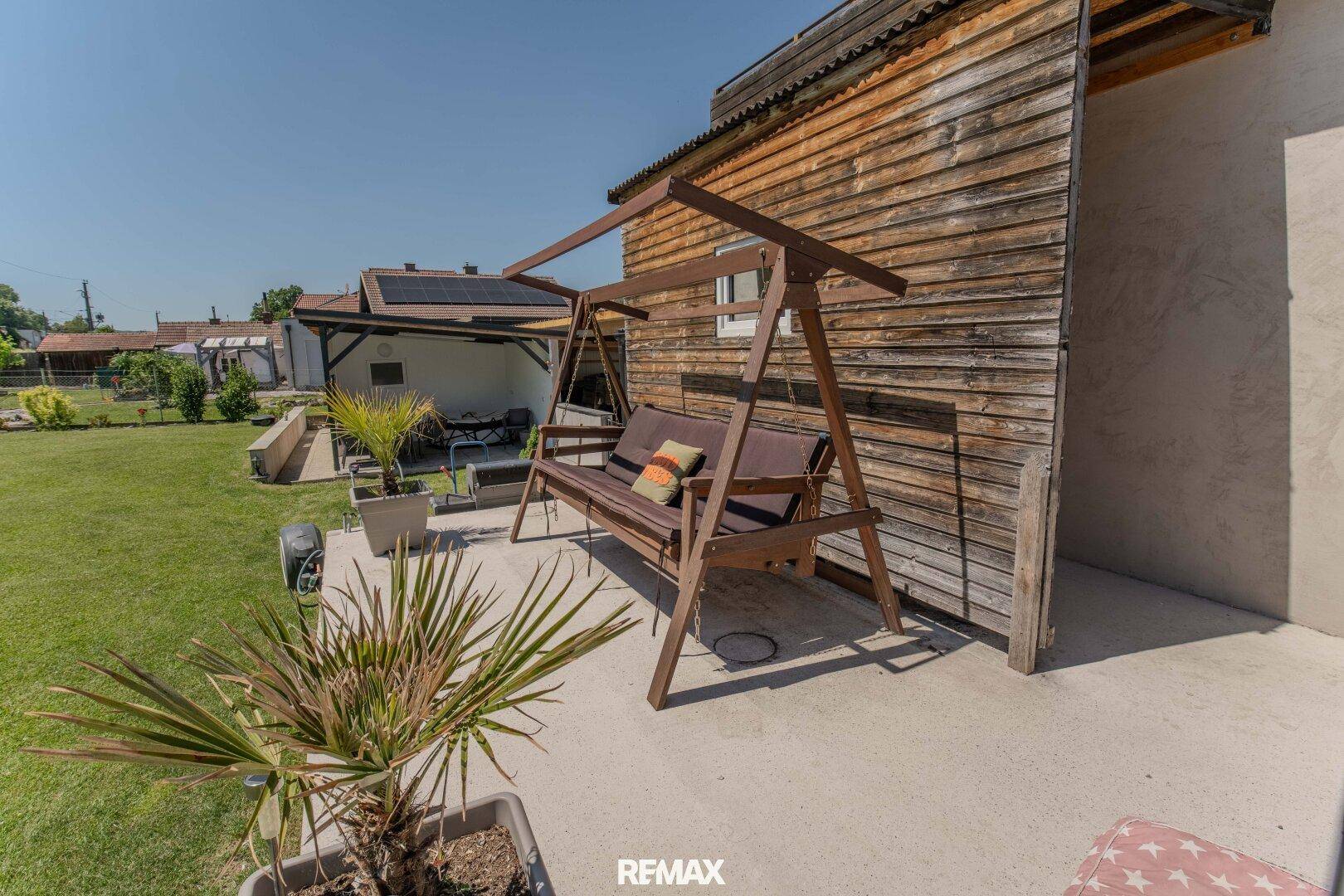 Gemütliche Terrasse mit Hollywoodschaukel und Pflanzen, angrenzend an die Holzfassade des Hauses.